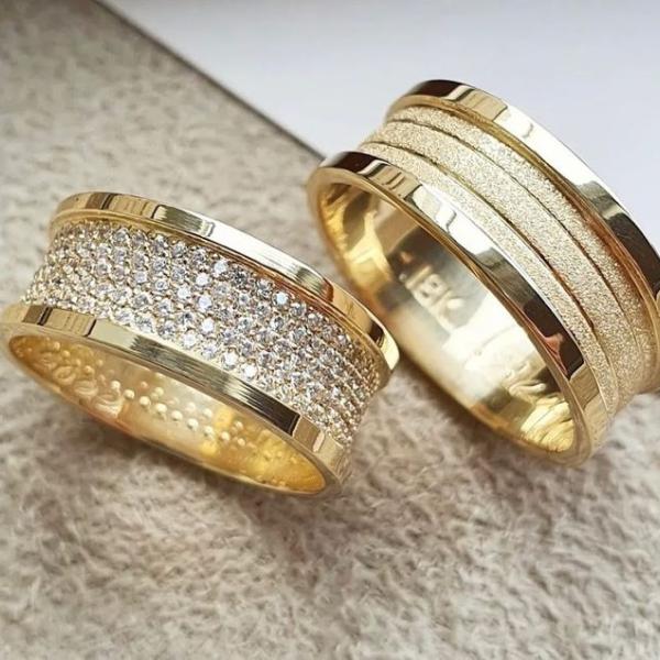 Modelo Carretel, com frisos na parte central, 9mm largura. No modelo 1: cravação de pedras (200 em média) em toda circunferência da aliança sendo na parte central. No modelo 2: diamantada na parte central em Ouro Amarelo - Tradicional 18k/750
